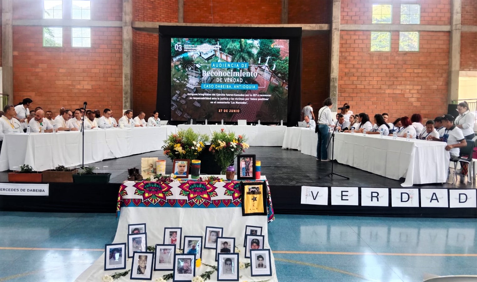 Mesa de participantes y mesa con fotos de victimas en audiencia por ejecuciones extrajudiciales
