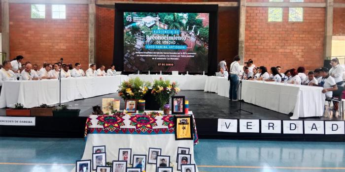 Mesa de participantes y mesa con fotos de victimas en audiencia por ejecuciones extrajudiciales